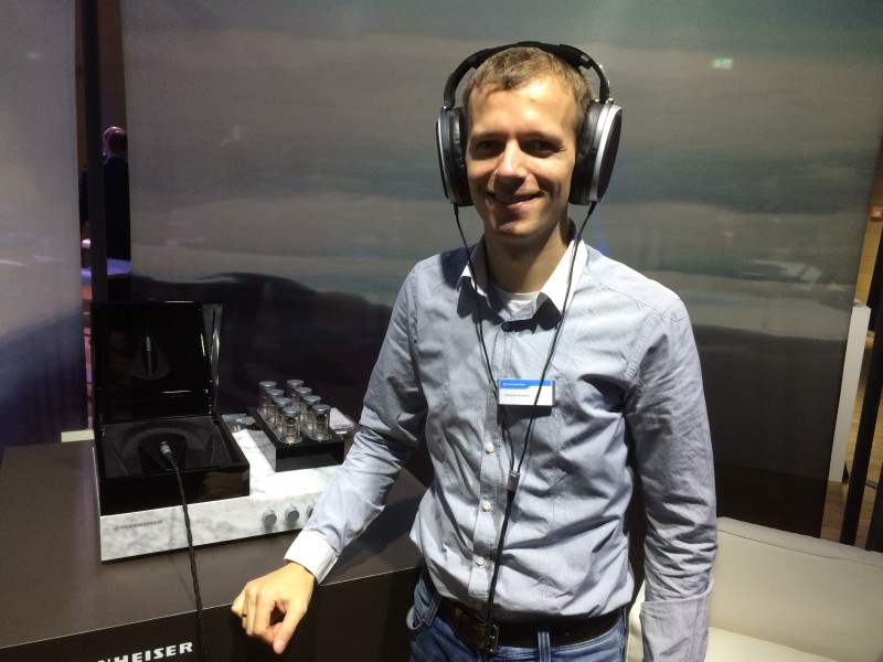 En man provar Sennheiser Orpheus hörlurar på en mässa med imponerande teknik och design i bakgrunden.