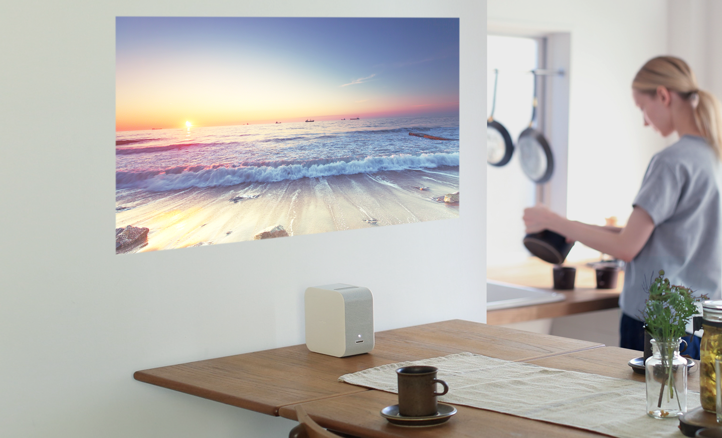 En kvinna brygger kaffe medan en liten Sony-projektor visar en strandbild på väggen.