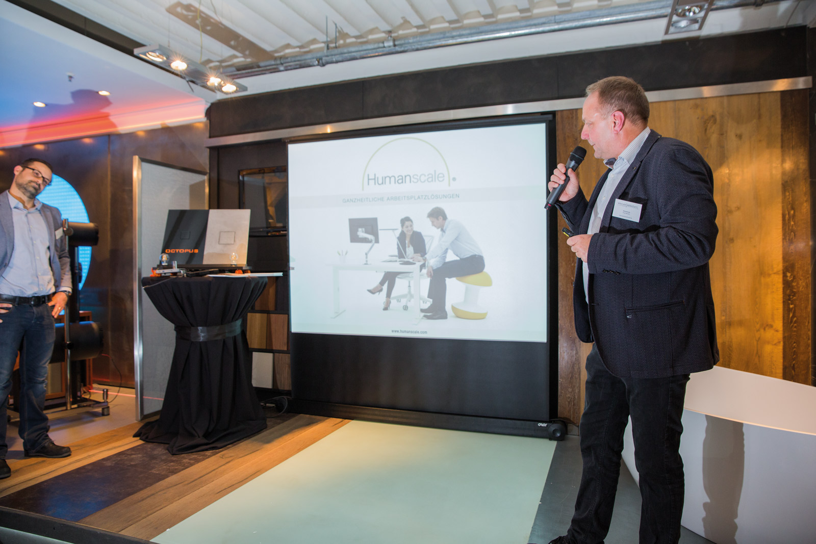 En föreläsare presenterar ergonomiska arbetsplatslösningar från Humanscale för en intresserad gäst i Hamburg.